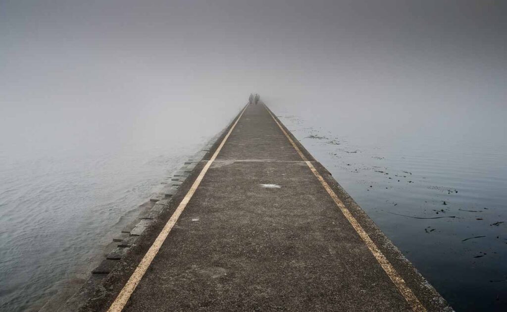 Foto de carretera que atraviesa el agua y desaparece en la niebla. Al fondo casi a punto de desaparecer, se atisban dos figuras humanas. Final de Vida.